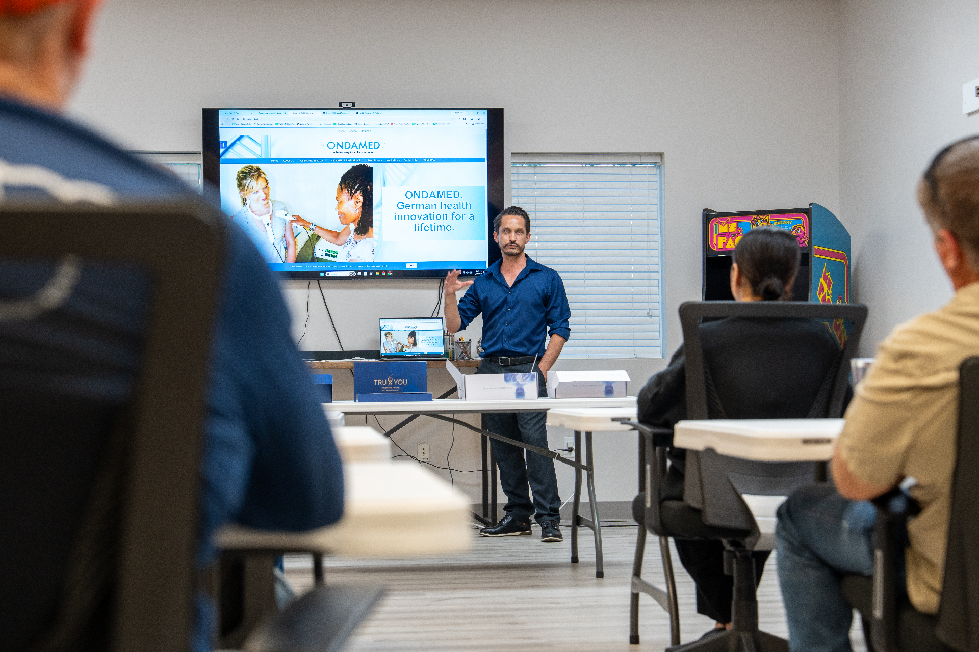 Speaker presenting to a seated audience in a bright room, with a large screen showing ONDAMED German health innovation and a Ms. Pac-Man arcade machine in the background.